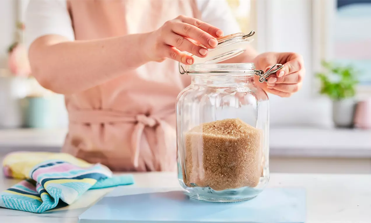 A large glass jar filled with coarse golden-brown sugar on a pastel blue surface, hands lifting the hinged lid, colourful towels nearby, AI-labeled image