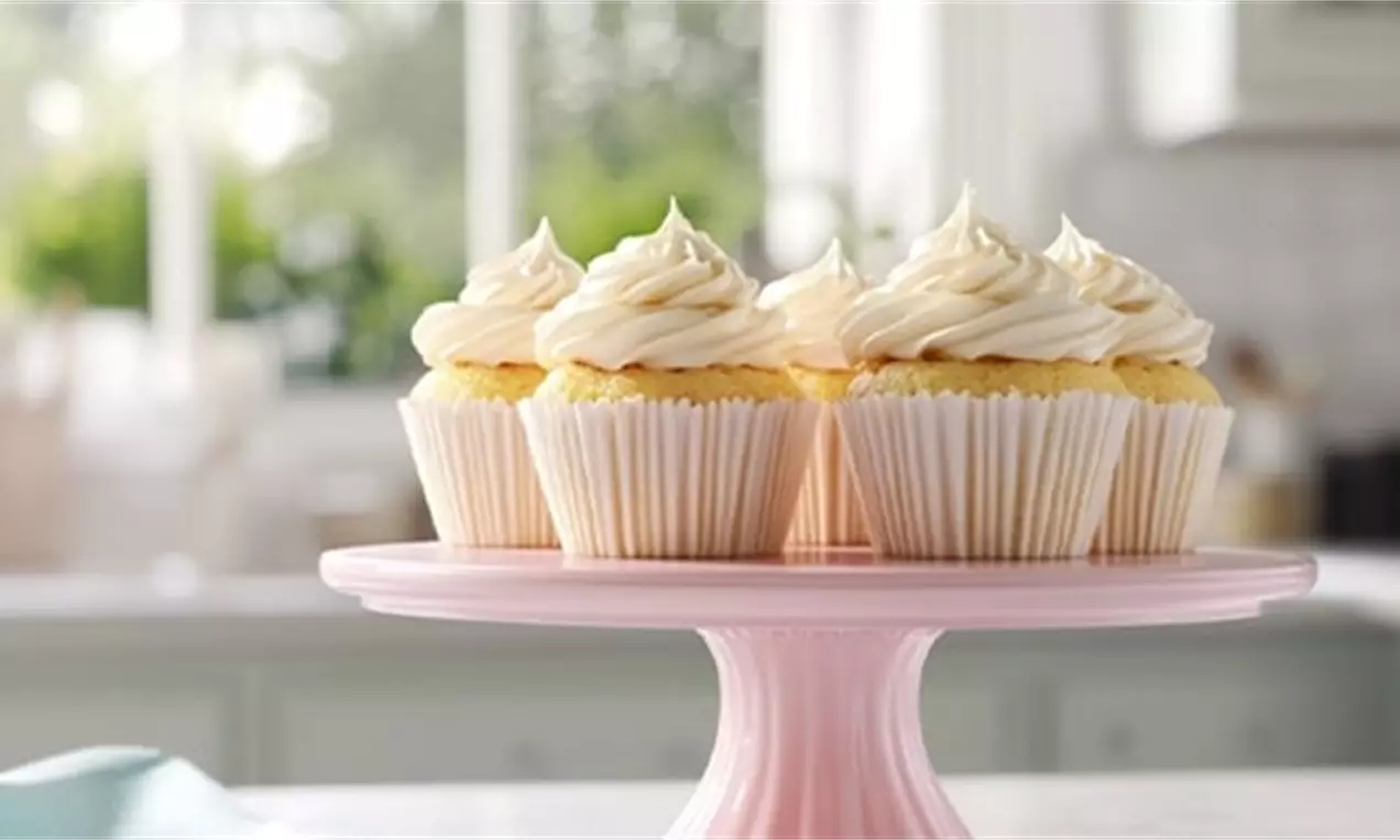 Golden yellow gluten-free vanilla cupcakes with swirled white frosting in ribbed paper cups arranged on a pastel pink cake stand, AI-labeled image