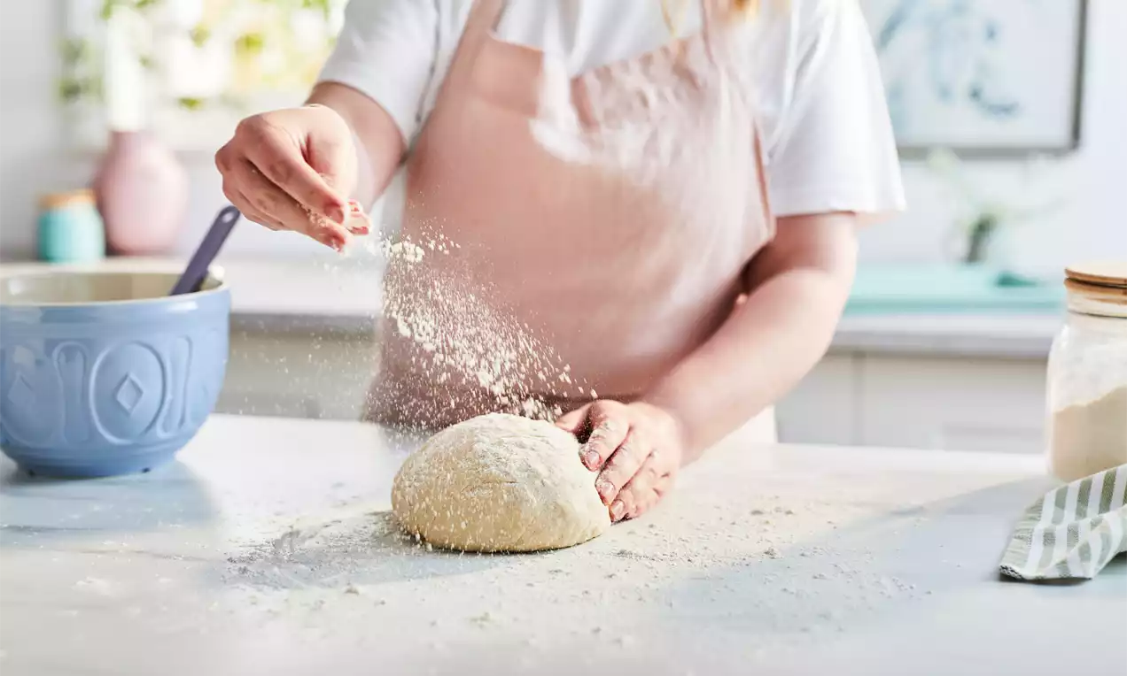 A ball of soft, pale dough on a floured white surface with a hand sprinkling flour, a blue bowl and jar in soft-focus background