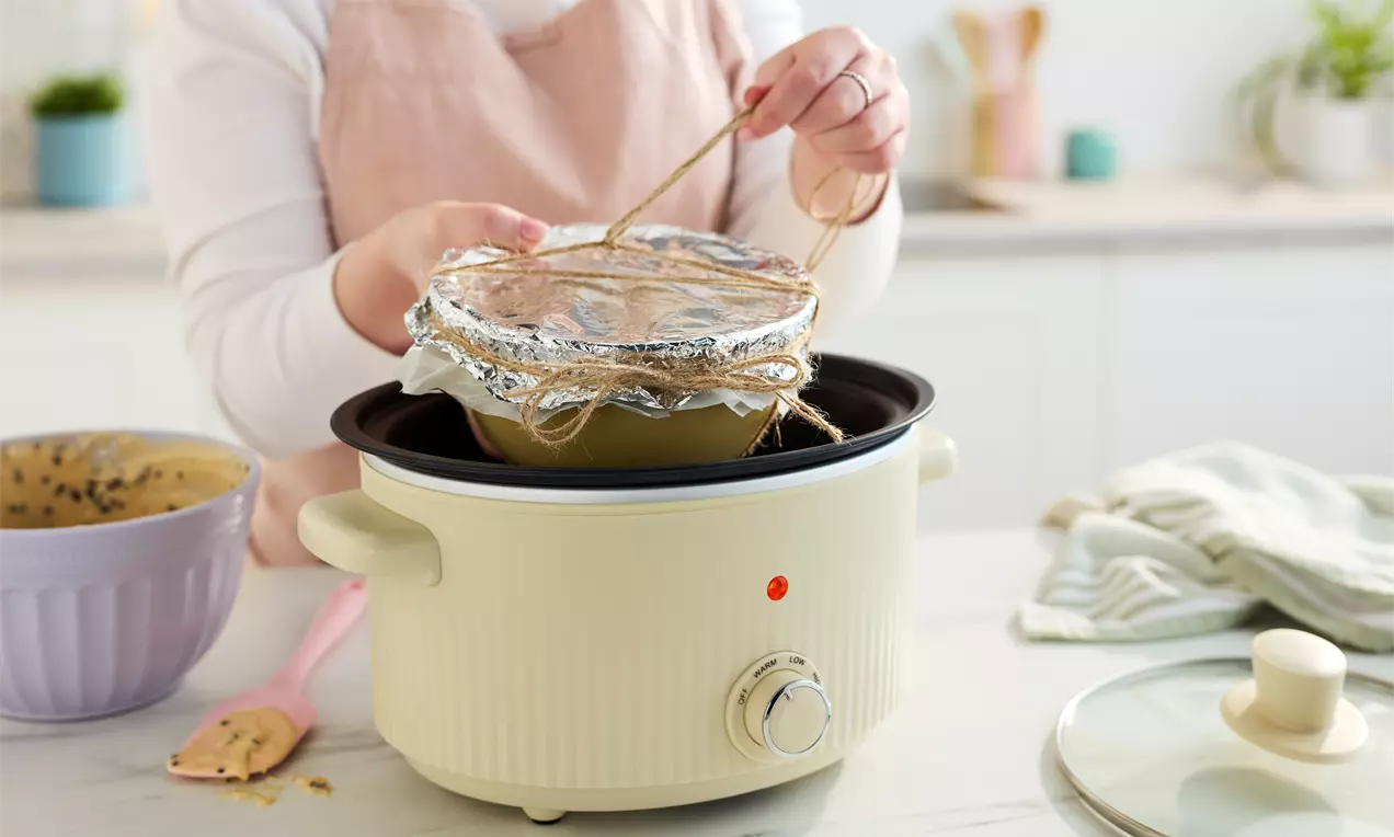 A beige slow cooker with a foil-covered bowl tied with twine inside, set on a white counter with pastel baking tools nearby