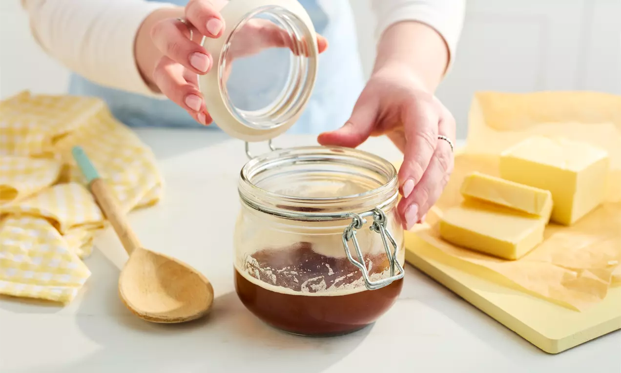A glass jar filled with warm brown butter, held open against a light kitchen backdrop with butter blocks, a wooden spoon, and yellow cloth