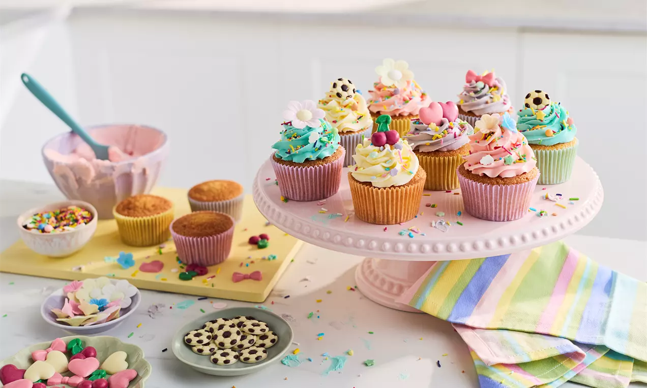 Colourful cupcakes with pastel frosting swirls, sprinkles, and shaped decorations on a pink cake stand, surrounded by baking items