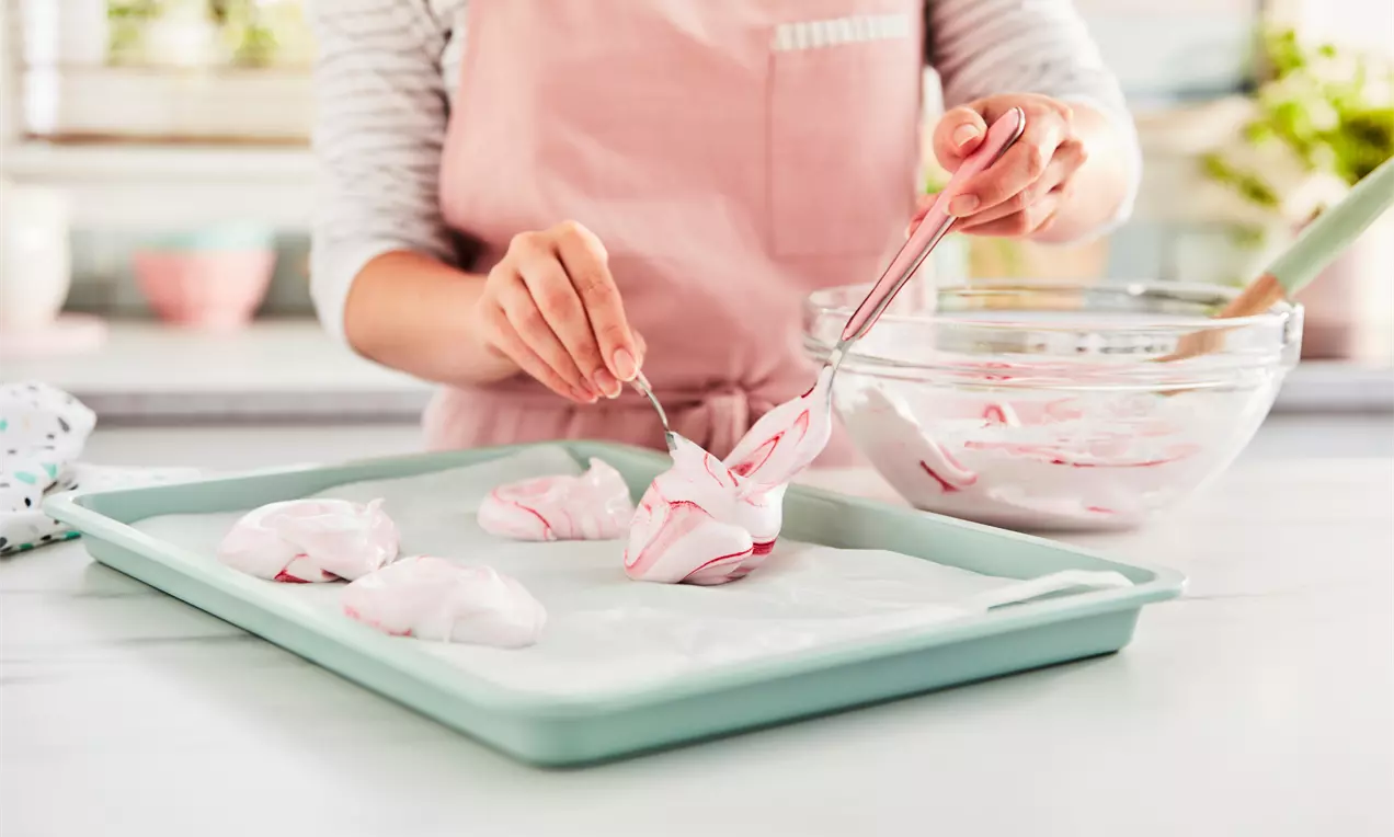 Soft pink and white swirled meringue dollops being shaped on a light green tray lined with parchment paper in a bright kitchen