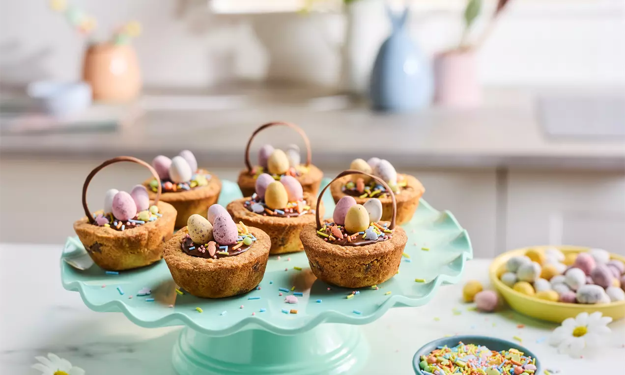Golden cookie nests with curved chocolate handles, filled with glossy chocolate, pastel candy eggs, and colourful sprinkles on a pale green stand