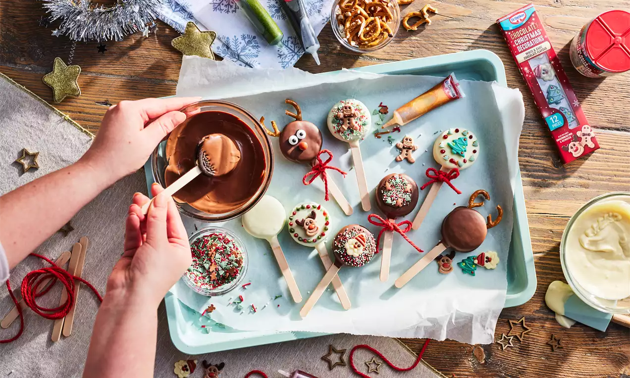 Oreos dipped in smooth chocolate, decorated with festive sprinkles and ribbon, arranged on wooden sticks on a light blue tray