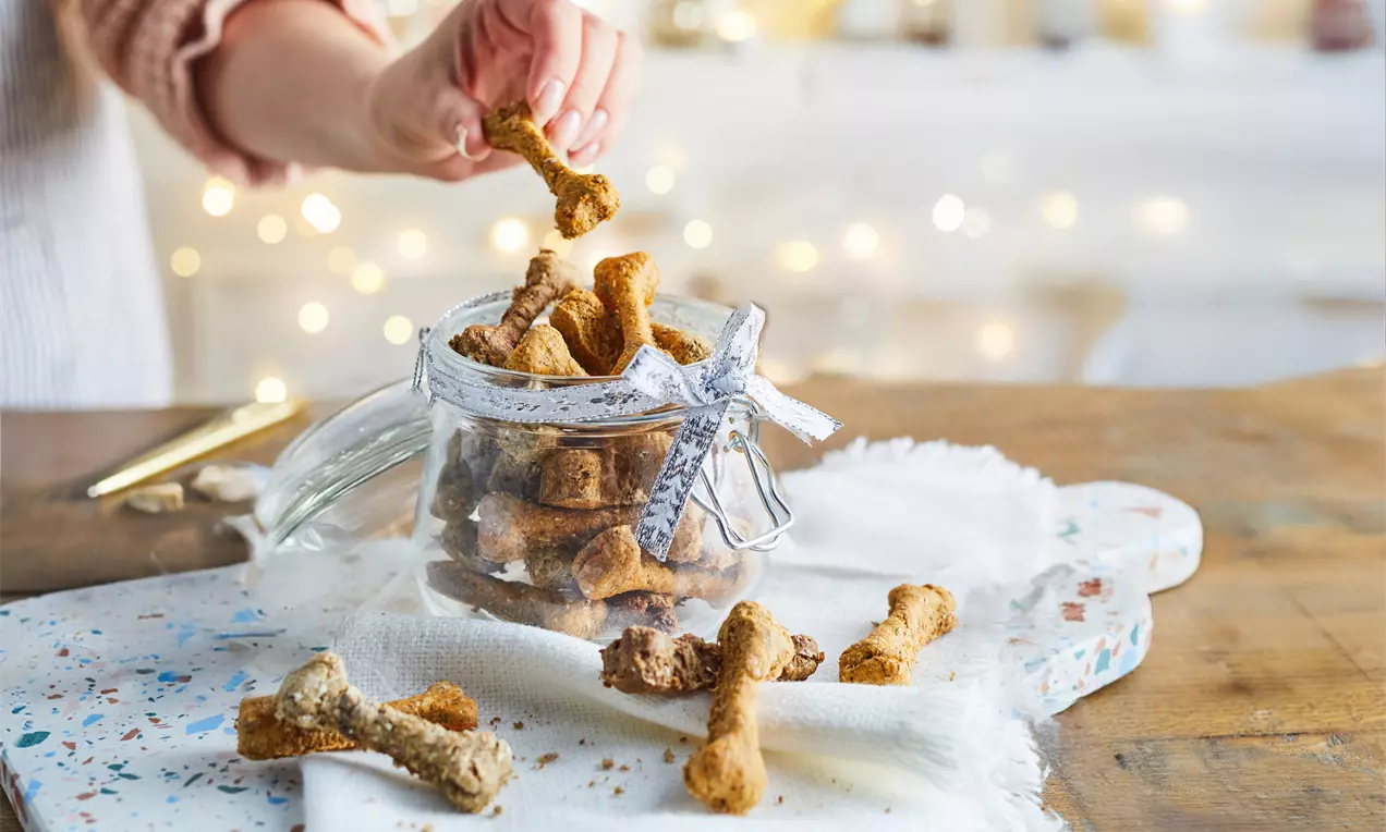 Golden-brown bone-shaped dog biscuits with a crumbly texture spilling from a glass jar tied with ribbon onto a white cloth, AI-labeled image