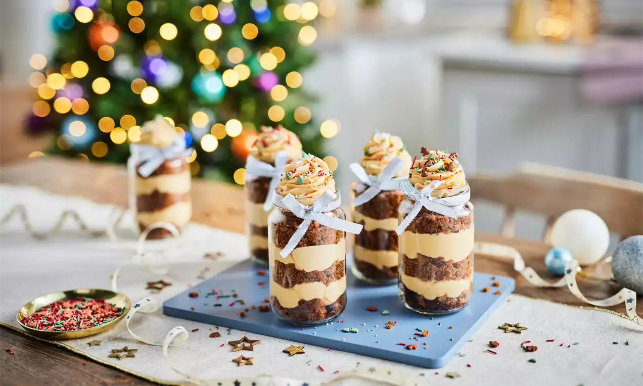 Layered desserts of caramel and chocolate sponge in glass jars tied with white ribbons, topped with cream swirls and sprinkles on a festive table