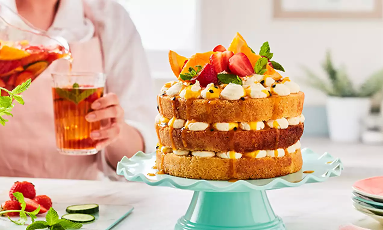 Three golden sponge layers with cream and fruit fillings, topped with fresh strawberries, orange slices, mint and served on a pale green stand