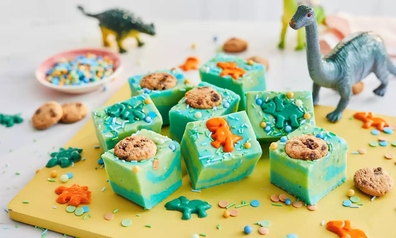 Vivid green and blue swirled fudge squares topped with tiny chocolate chip cookies, orange and green dinosaur shapes, and sprinkles on a yellow board