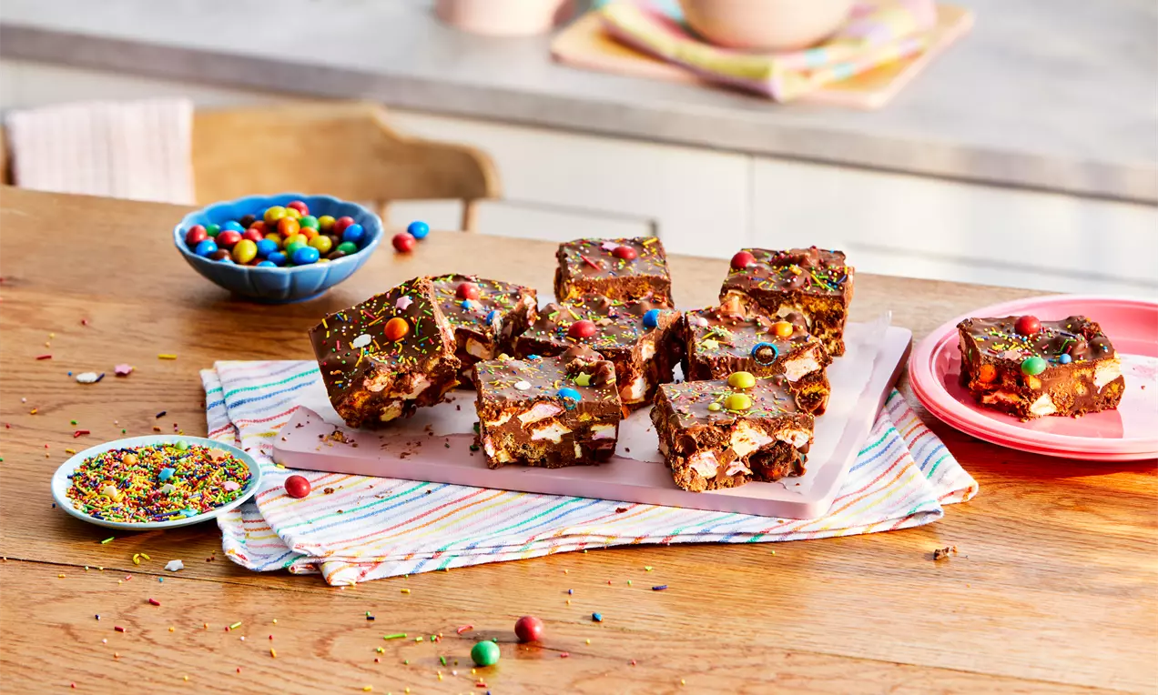 Chocolate rocky road squares sprinkled with colourful candy pieces and small marshmallows, arranged on a pink tray on a striped cloth