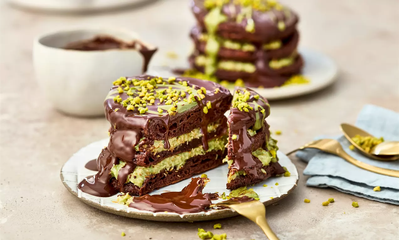 Layered chocolate pancakes with glossy melted chocolate, green pistachio cream, and crushed pistachios on a white plate