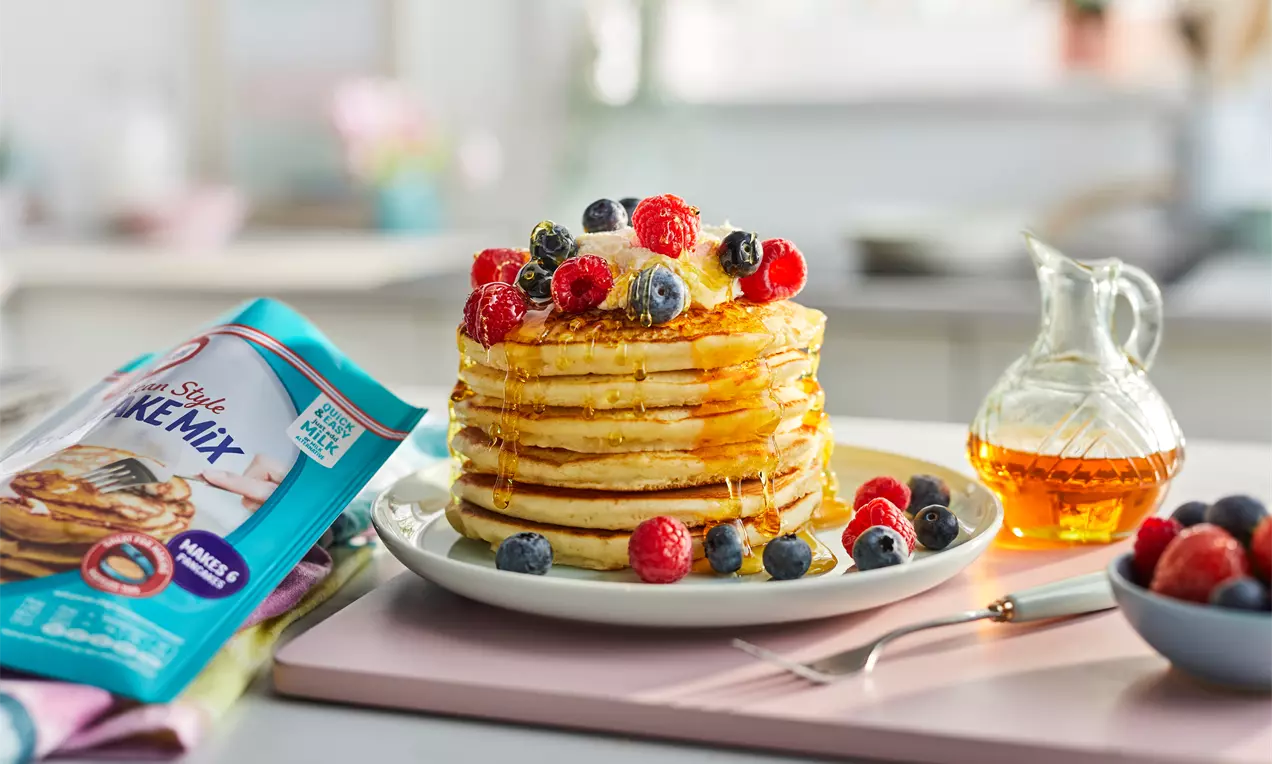 A stacked tower of golden brown American pancakes drizzled with syrup, topped with fresh raspberries, blueberries, and butter on a white plate