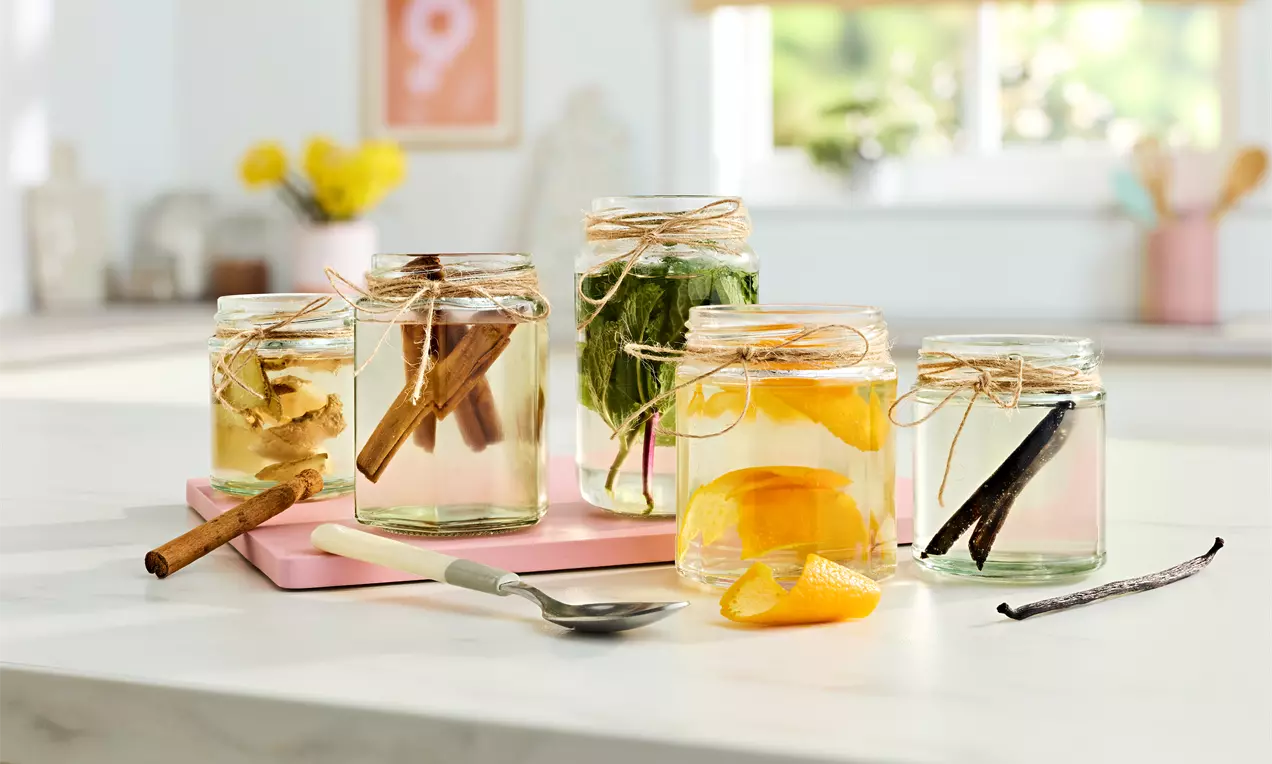 Clear glass jars filled with transparent liquid, infused with cinnamon sticks, herbs, ginger, orange slices, and vanilla beans on a countertop