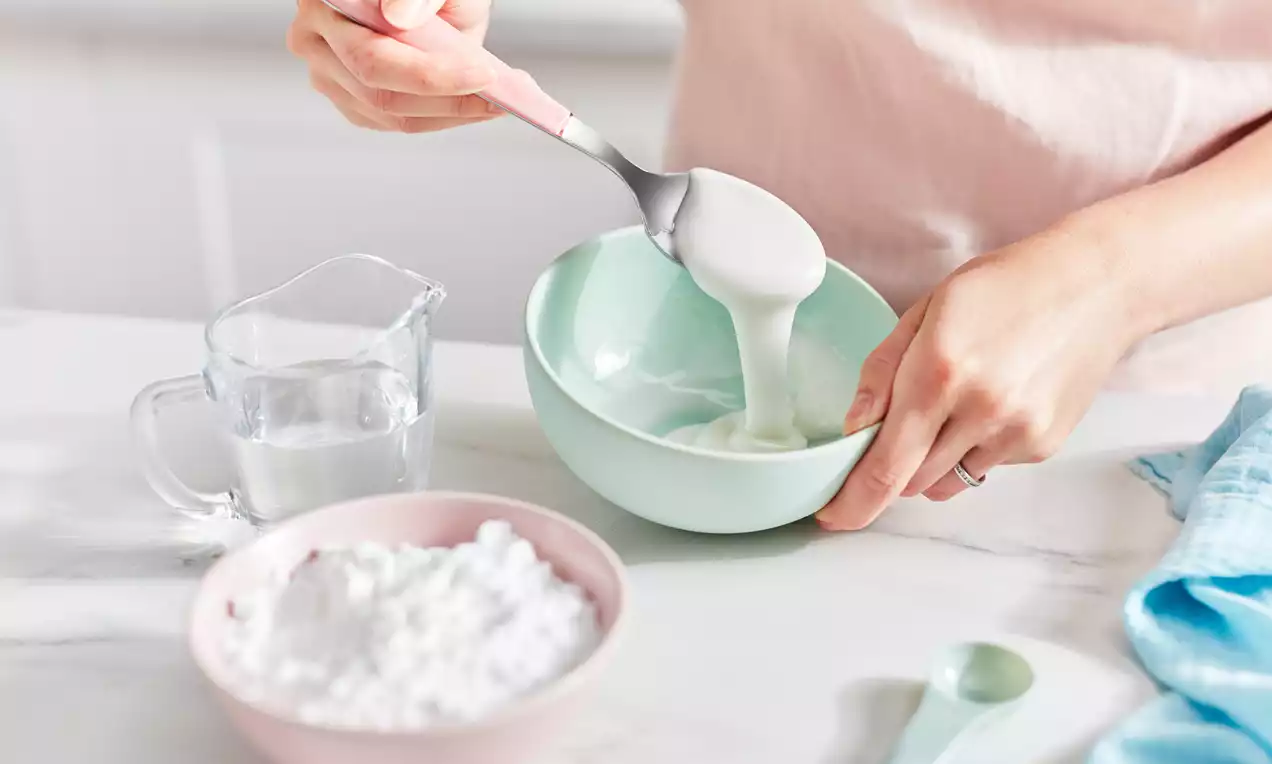 Smooth, glossy white icing drips from a spoon into a pastel green bowl beside a pink bowl of sugar, a glass jug, and a pale blue cloth
