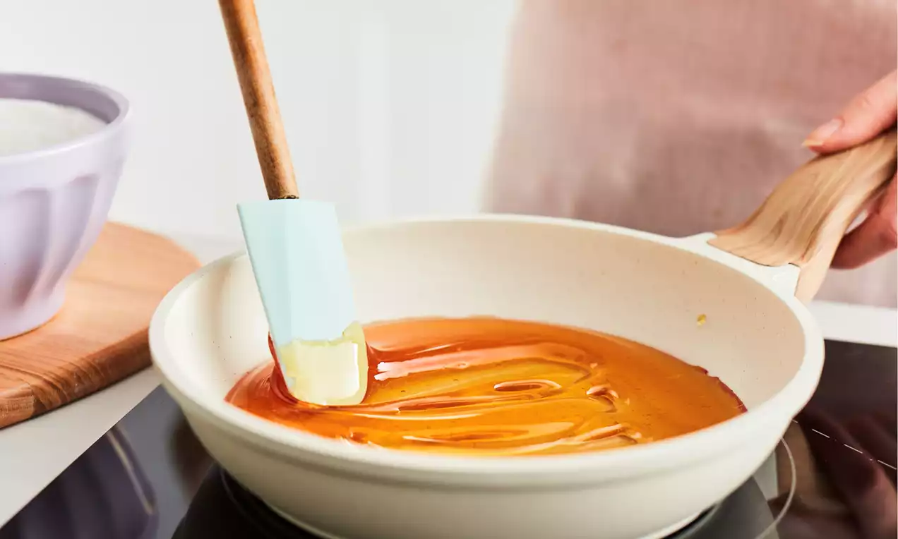 Golden caramel being stirred smoothly with a light blue spatula in a round cream-coloured pan on a black stovetop, AI-labeled image