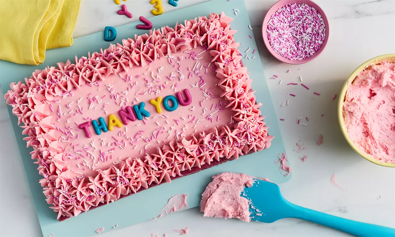 Rectangular pink traybake with piped star edges, colourful “THANK YOU” letters in the centre, and pink sprinkles on a light blue board