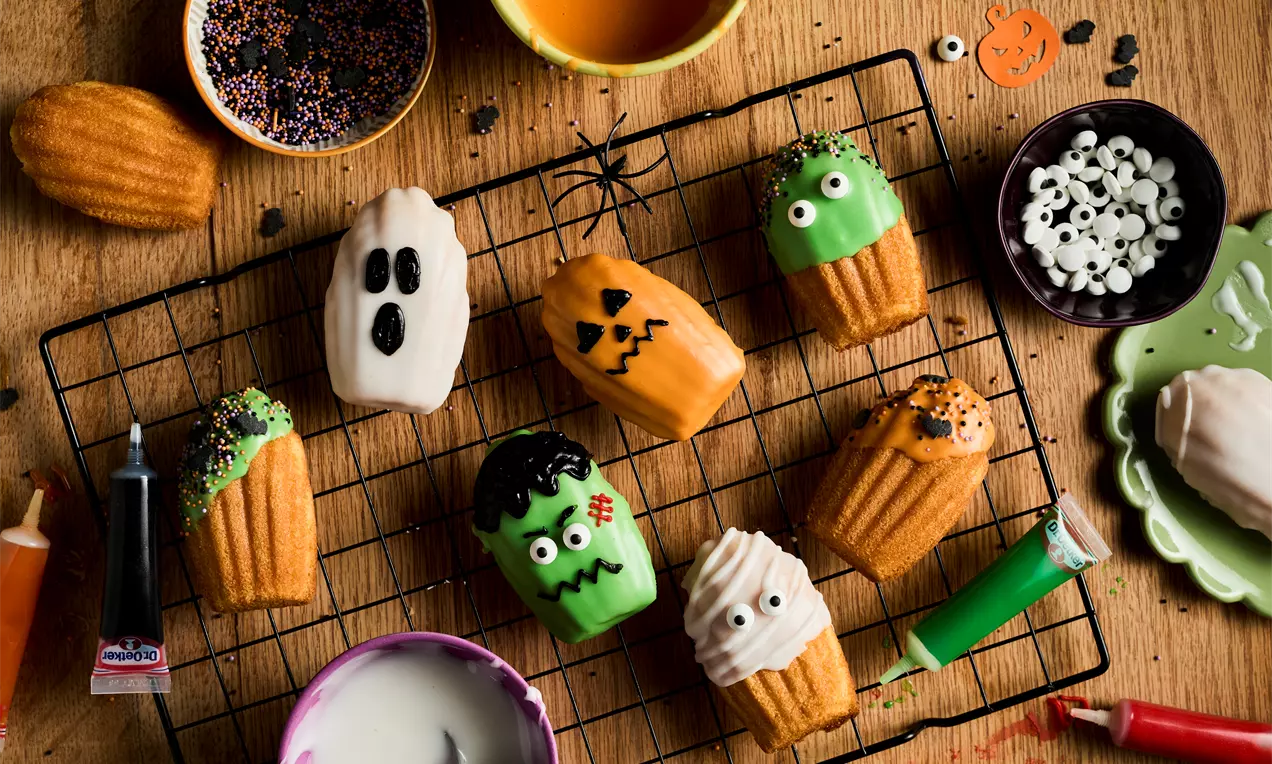 Madeleines in Halloween designs featuring orange pumpkins, white ghosts, and green monsters with icing and candy eyes on a black cooling rack
