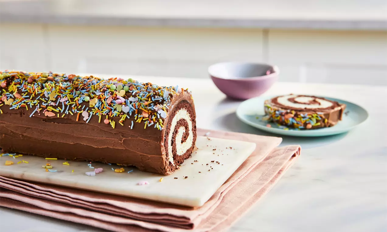 Chocolate Yule log with a spiral cream filling, smooth frosting, colourful sprinkles, served on a marble board with slices on plates, AI-labeled image