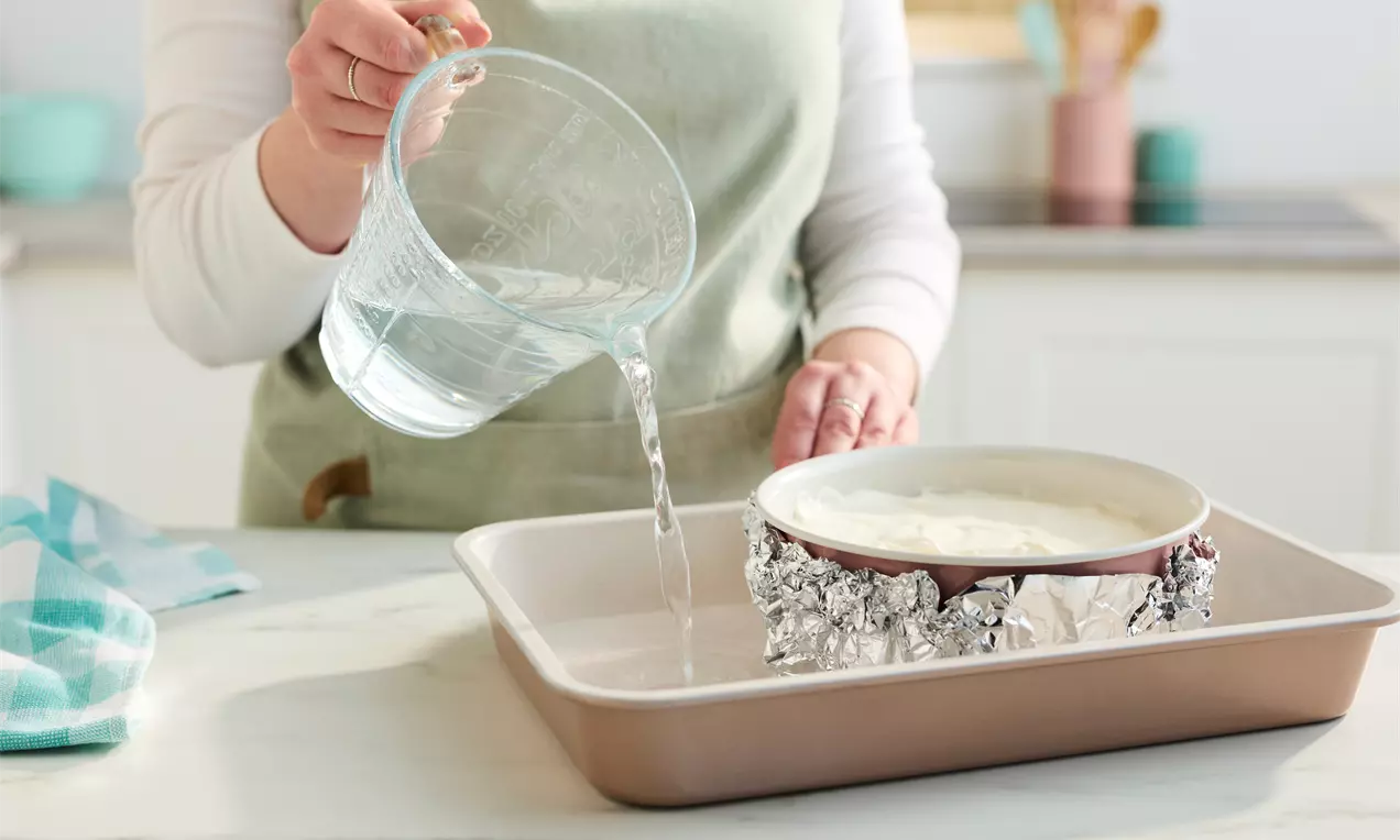 A creamy off-white cheesecake in a foil-wrapped pan placed in a beige baking dish while water is poured from a clear glass jug