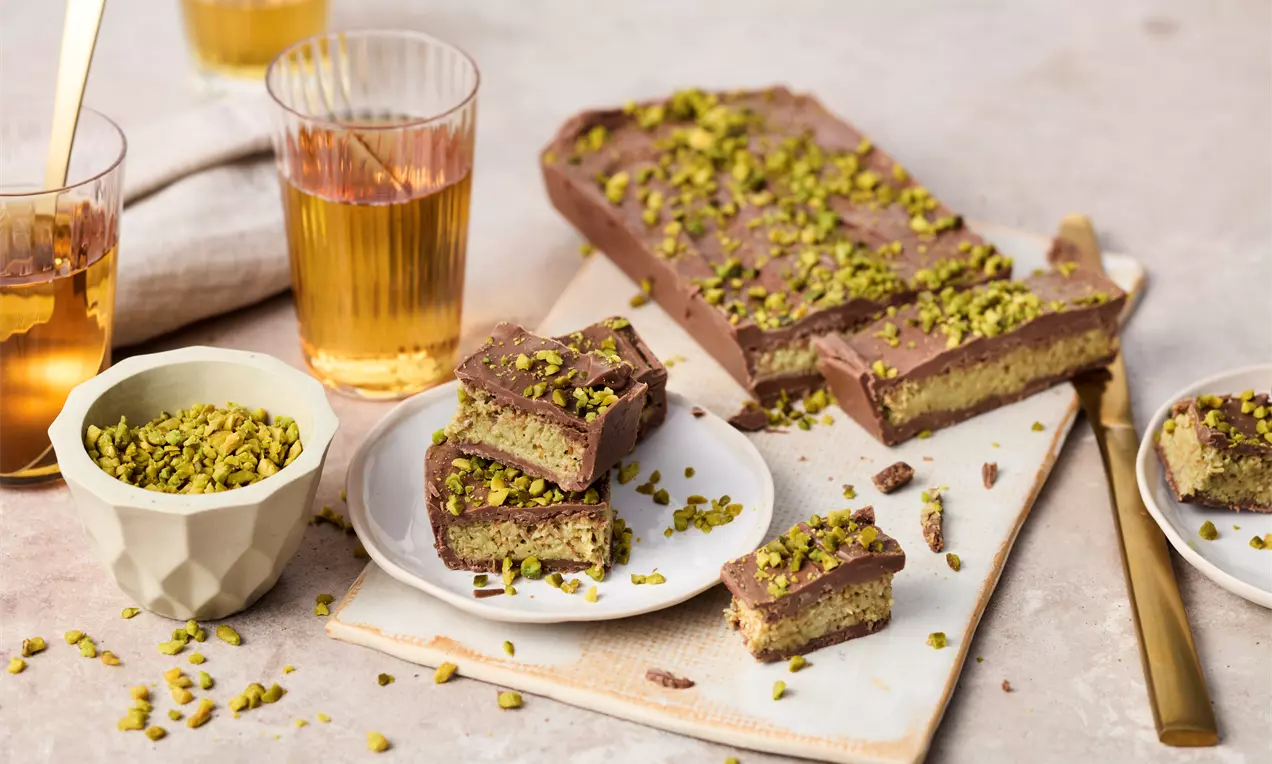Rectangular chocolate squares layered with golden pistachio filling, topped with crushed green pistachios on white plates