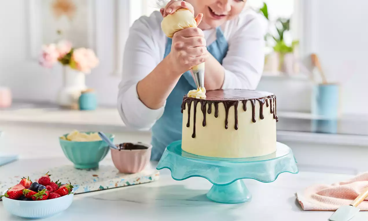 A round cake with smooth pale cream frosting and glossy dark chocolate drips on a blue glass stand, being decorated with piped cream.