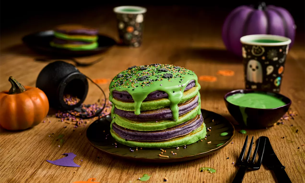 A stack of green and purple pancakes layered neatly, topped with dripping green glaze and sprinkles on a dark plate surrounded by Halloween-themed decorations