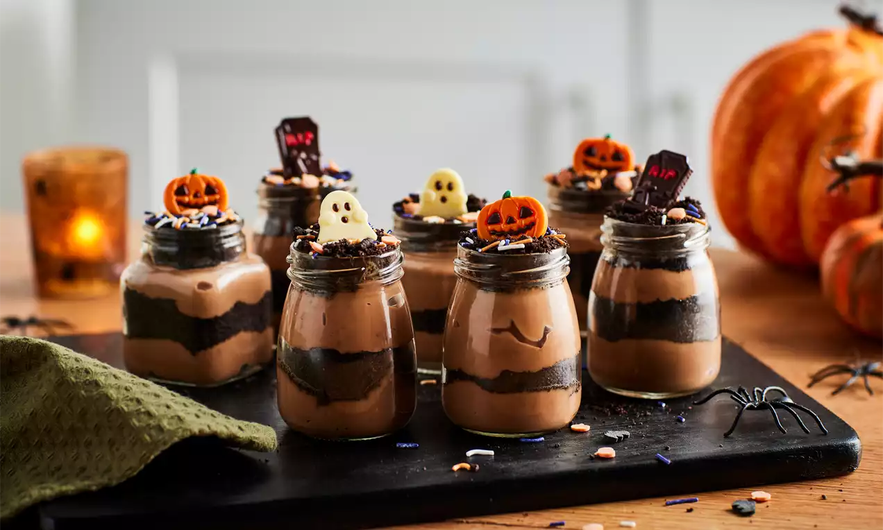 Glass jars layered with smooth chocolate mousse and dark cookie crumbs, topped with Halloween decorations and sprinkles on a slate board