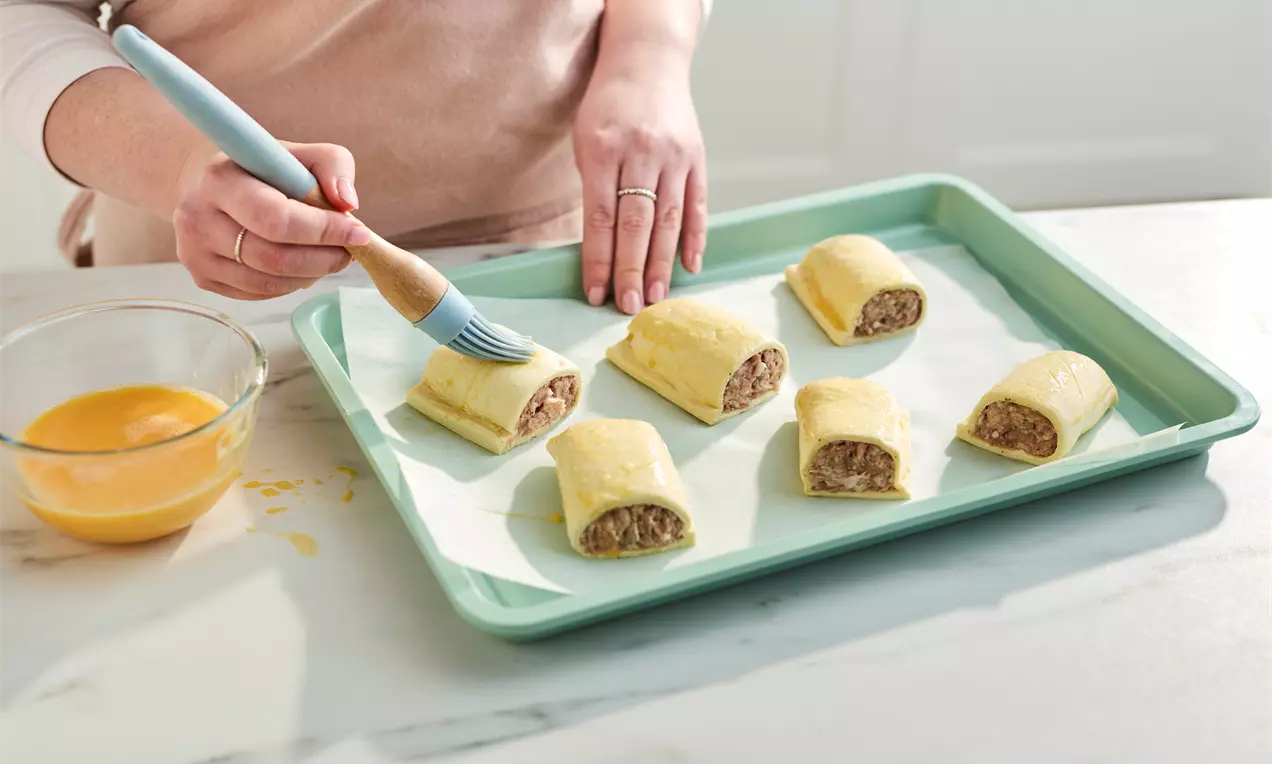Pale golden pastry rectangles filled with minced meat on a mint-green tray, brushed with glossy egg wash using a blue-handled brush